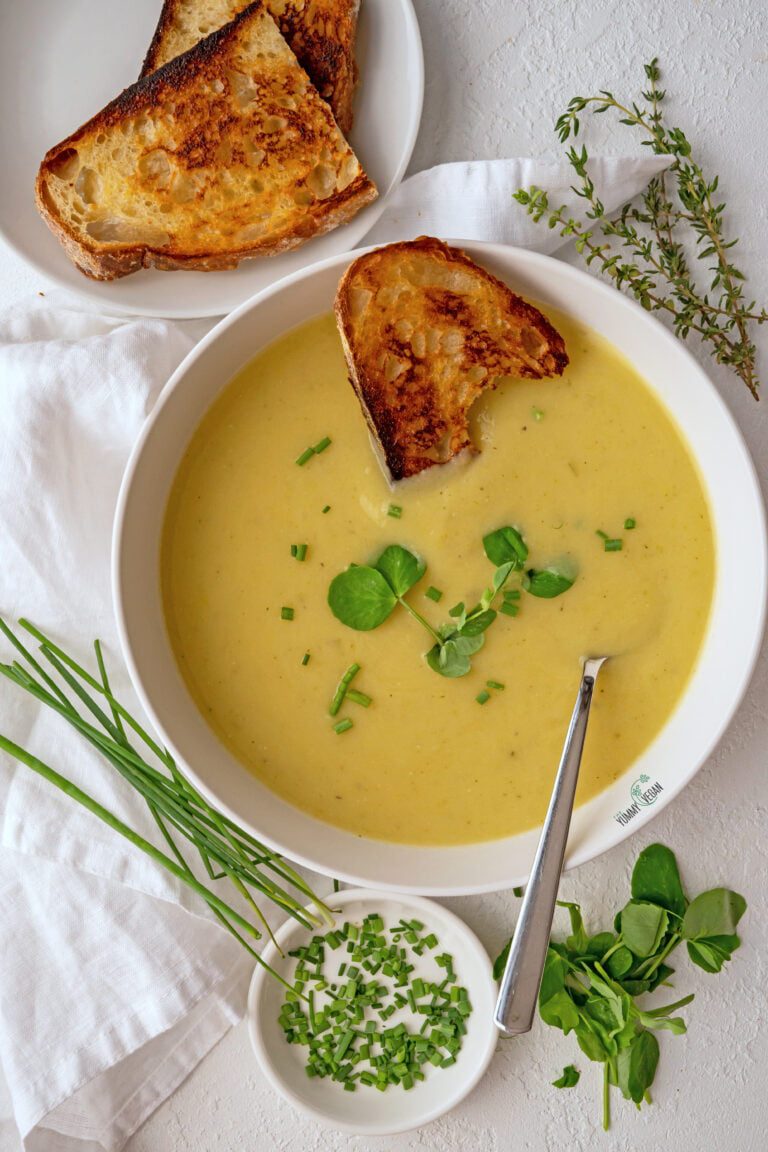Vegan Potato Leek Soup in a bowl with a spoon, a toasted slice of bread dunked in the soup and more bread on a plate on the side, herbs all around
