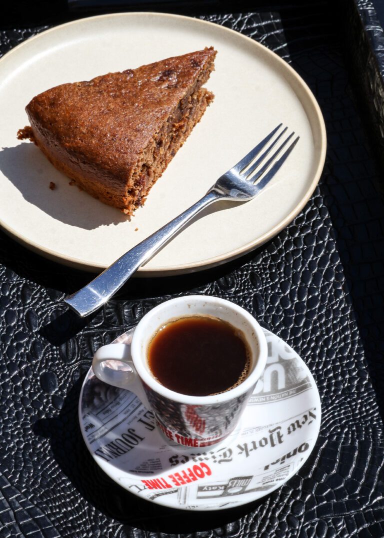 A piece of Tahini Date Cake on a plate with a fork next to a cup of coffee